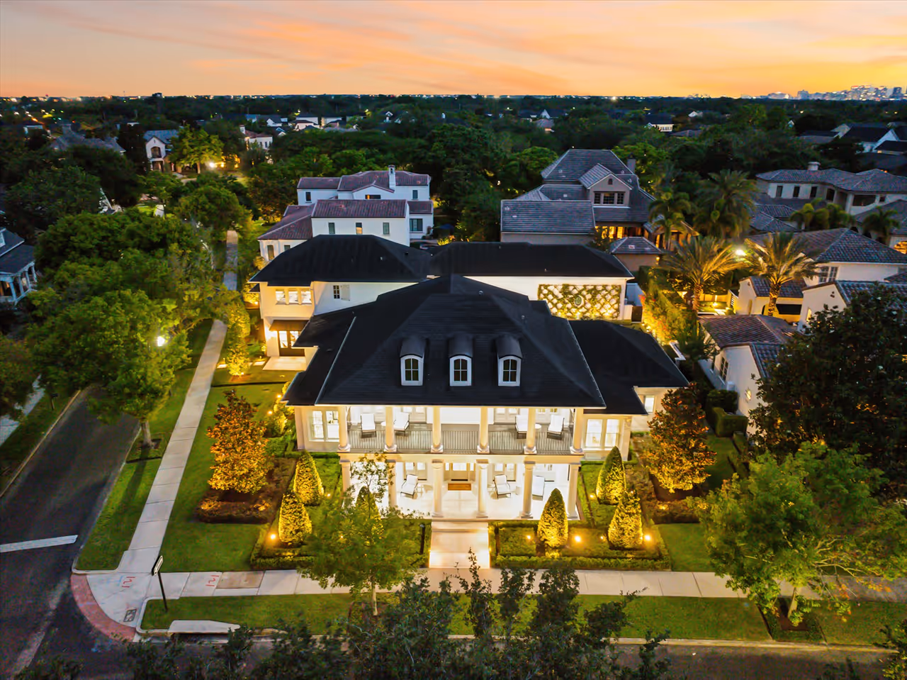 Aerial view of a large, illuminated two-story house with a dark roof and symmetrical columns, surrounded by landscaped trees and neighboring homes at dusk.