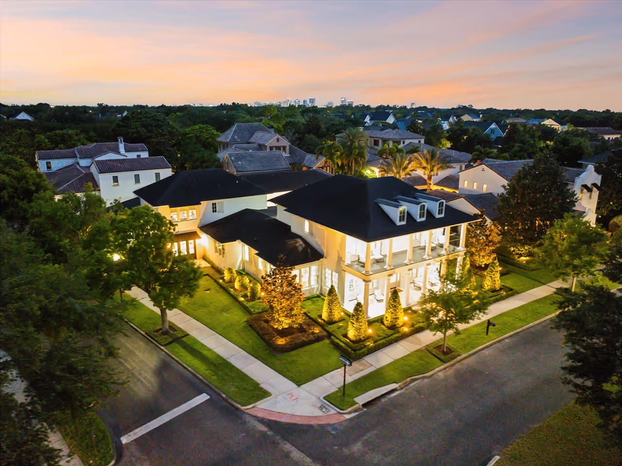 A large, well-lit two-story house with white walls and black roof on a corner lot at dusk, surrounded by trees and other homes.