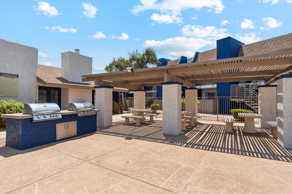 Outdoor communal area with two stainless steel grills on a blue counter and shaded picnic tables under a pergola at an apartment complex.