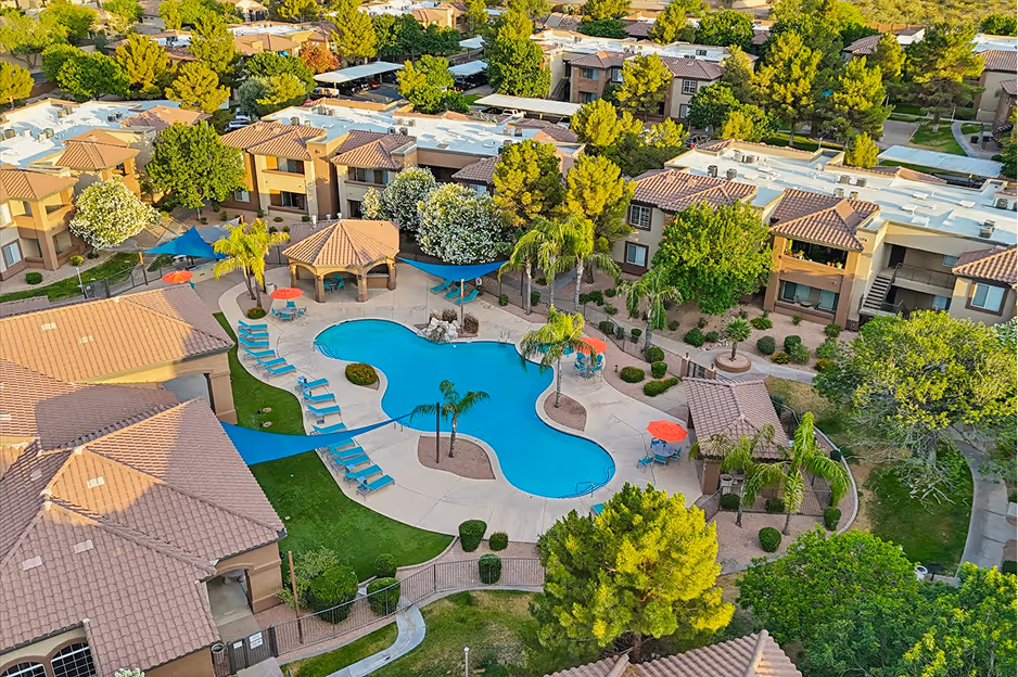 Aerial view of a residential community with a kidney-shaped swimming pool surrounded by lounge chairs, umbrellas, palm trees, and beige stucco buildings with tiled roofs.