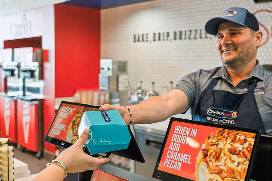 Smiling Cinnabon employee handing a teal Cinnabon box to a customer inside a store with a sign that reads 'When in doubt add caramel pecan.'