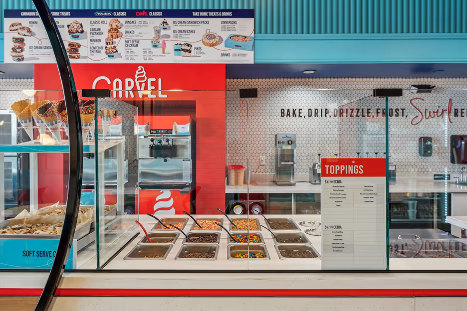 Ice cream toppings counter with various colorful toppings in metal containers and cones displayed on the left side inside a Carvel store.