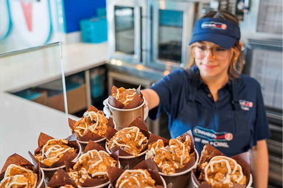 Person wearing glasses and a blue uniform holding a tray of cinnamon rolls topped with icing in a bakery or café setting.