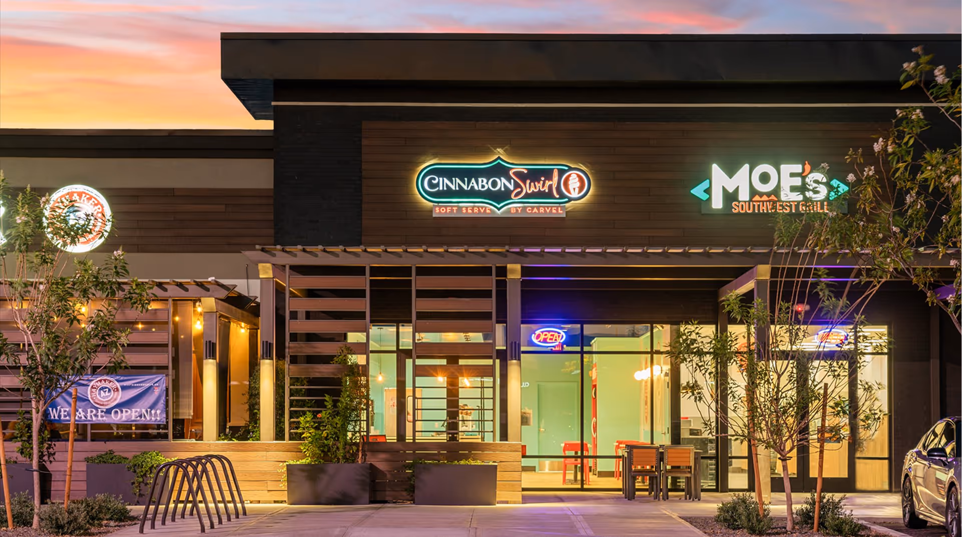 Exterior view of a shopping plaza at sunset featuring storefronts of Cinnabon Swirl and Moe's Southwest Grill with outdoor seating and trees.