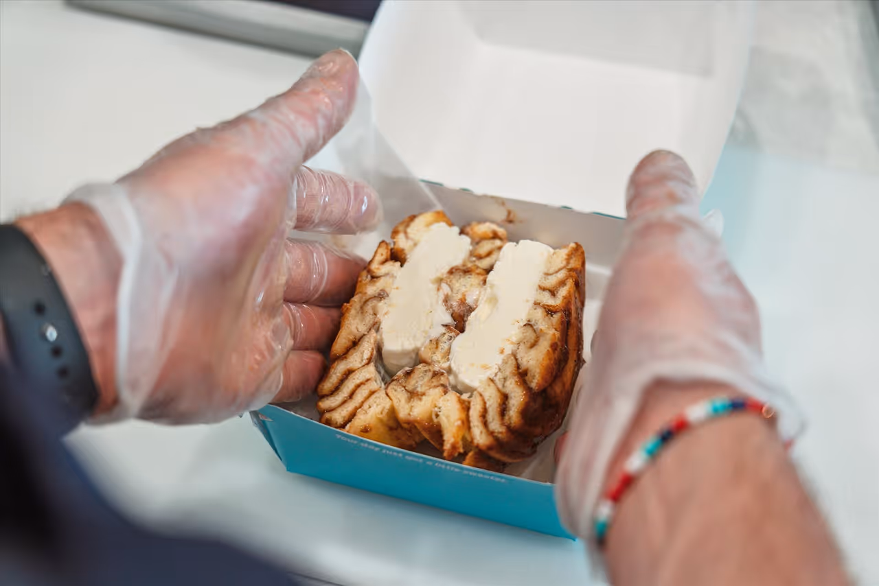 Hands wearing transparent gloves holding an open blue box containing a cinnamon roll with cream cheese frosting.