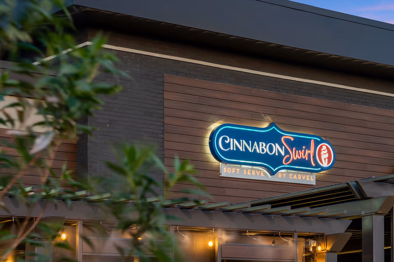 Illuminated Cinnabon Swirl sign on a wooden and brick building exterior at dusk.