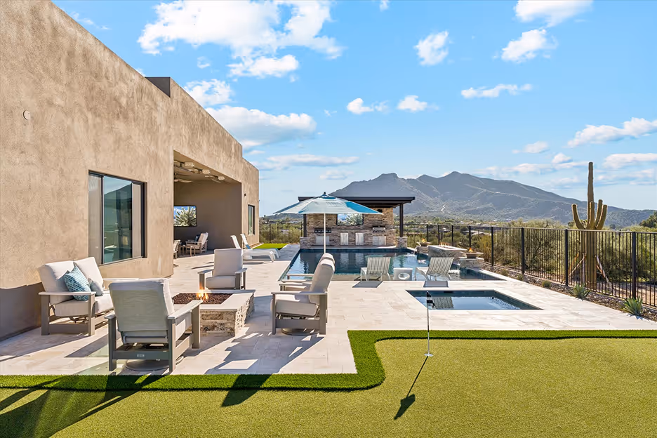 Modern backyard with patio seating around a fire pit, pool with lounge chairs and umbrella, putting green, and mountain views under a blue sky.