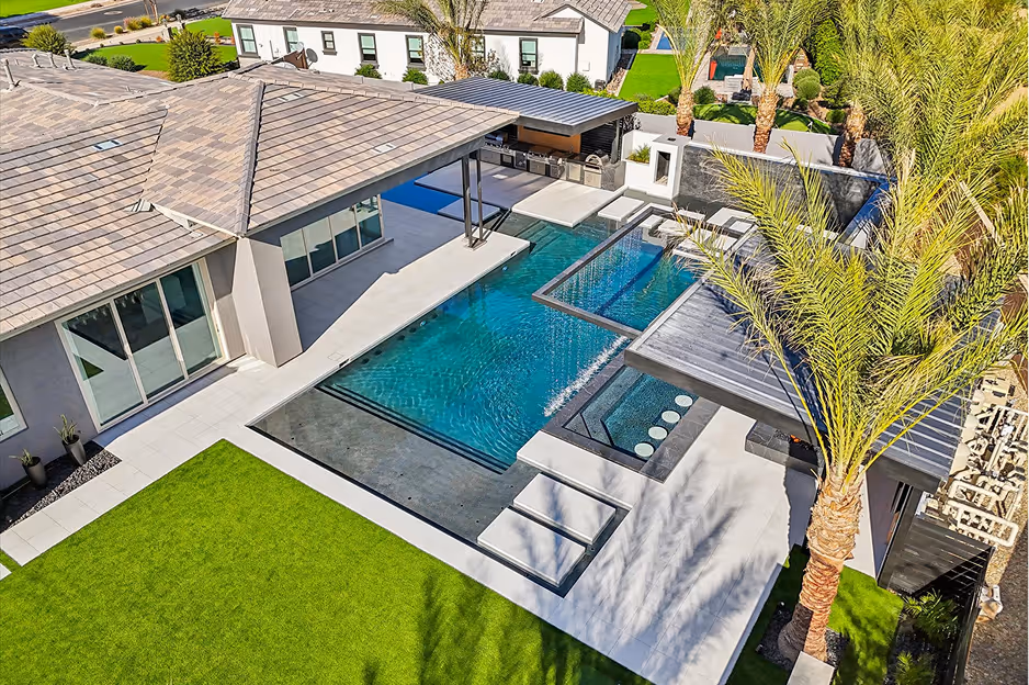 Modern backyard with a rectangular swimming pool, integrated hot tub, outdoor kitchen, and surrounding palm trees.