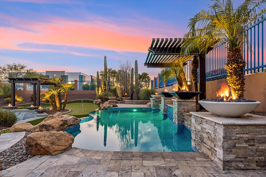 Backyard with a swimming pool bordered by stone fire pits, palm trees, and a putting green under a colorful sunset sky.