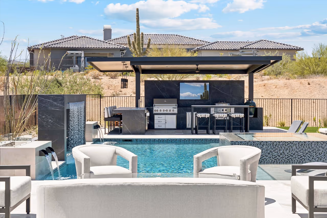 Modern outdoor kitchen and seating area with a covered patio, barbecue grills, bar stools, and a swimming pool with water features.