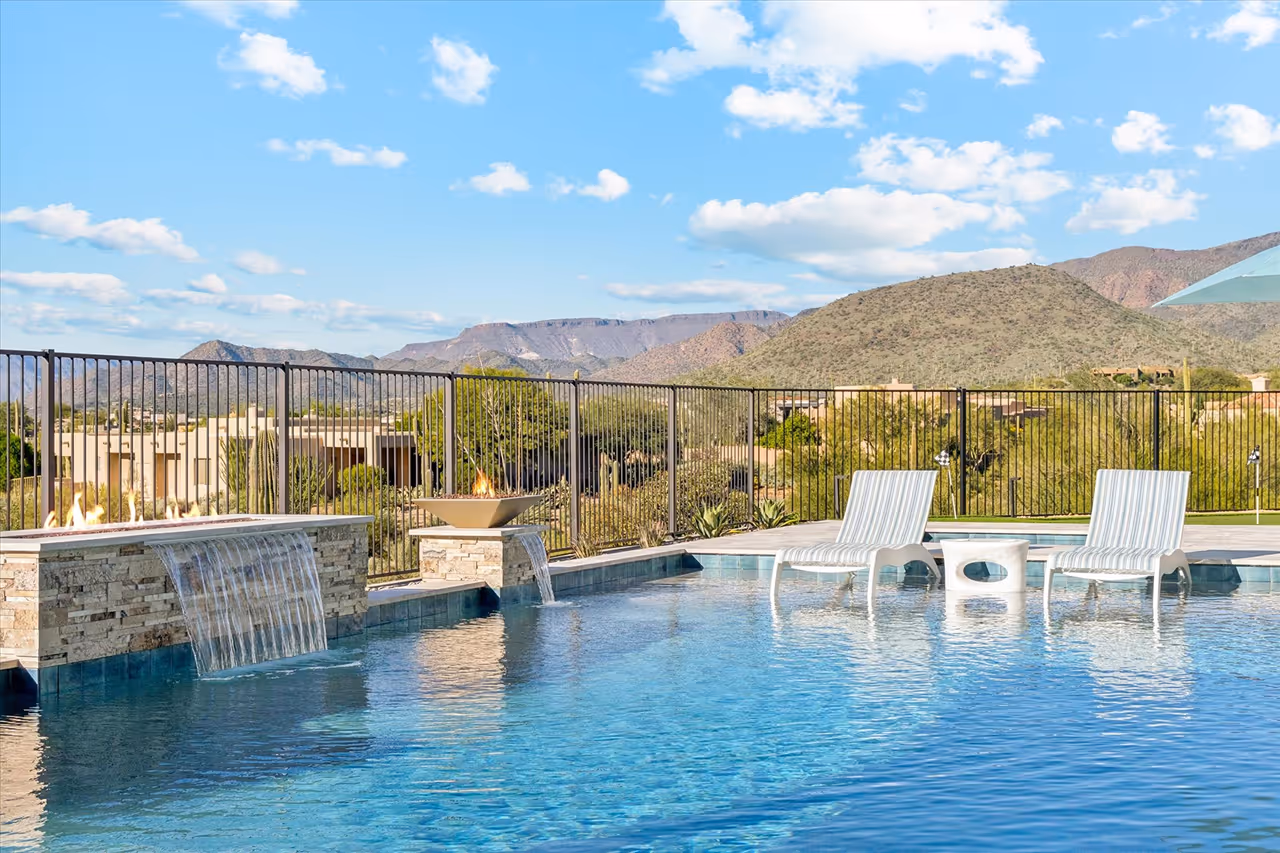 Swimming pool with two white lounge chairs and small table on the deck under a clear blue sky with mountains in the background.