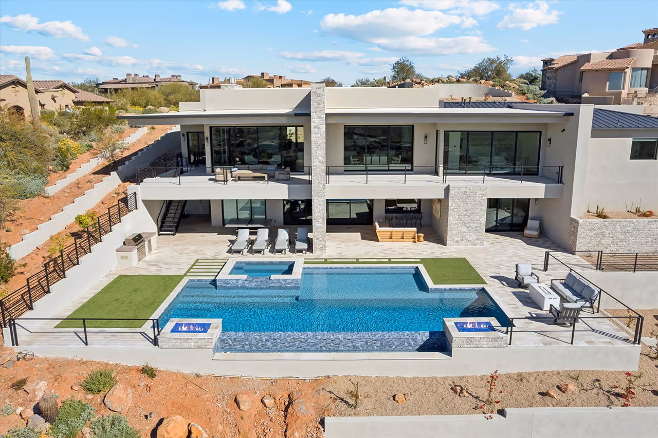 Modern two-story house with large glass windows, balcony, outdoor lounge areas, lawn patches, and a rectangular swimming pool with fire features.