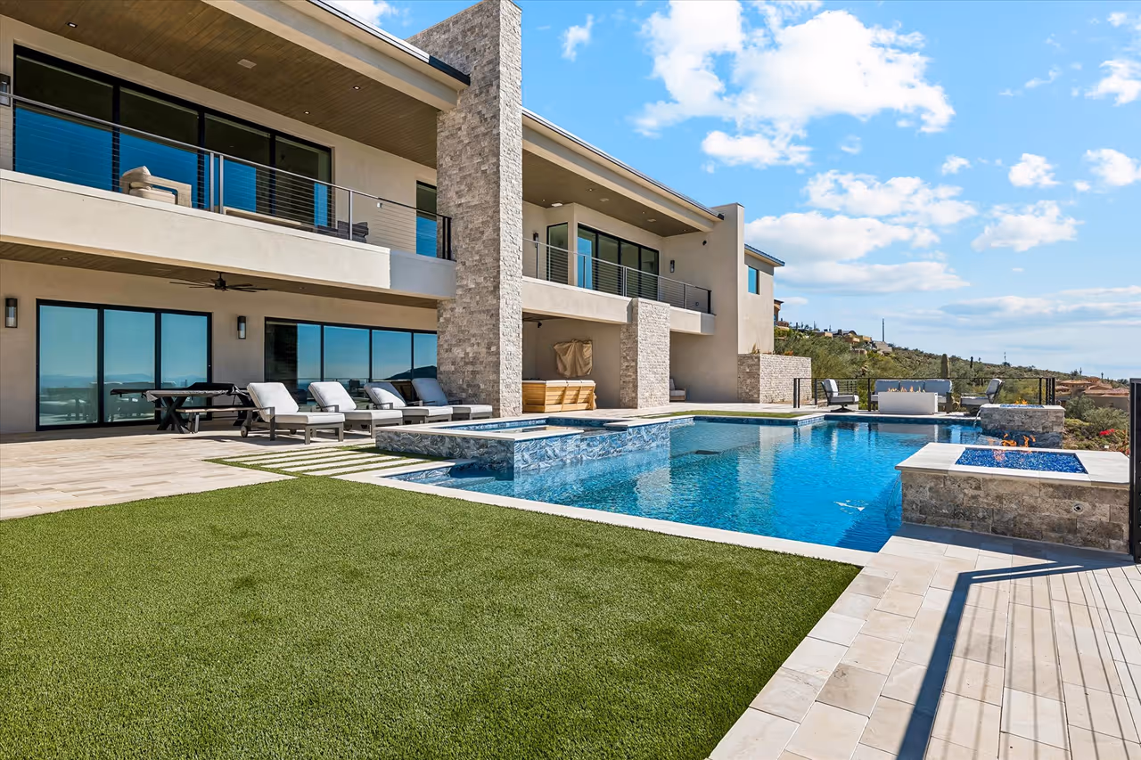 Modern house with large balcony, outdoor lounge chairs, swimming pool, and fire pit under a blue sky.