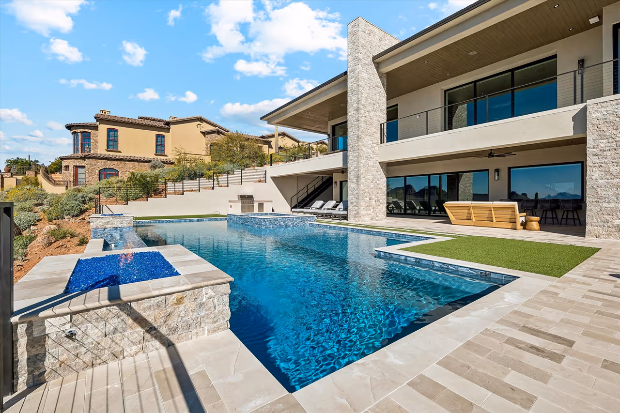 Modern house with large glass windows overlooking a rectangular swimming pool with a built-in fire feature and lounge chairs nearby.