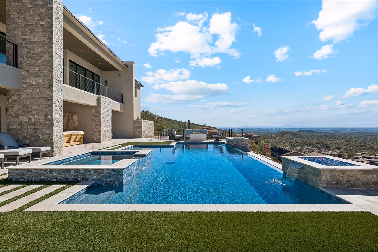 Modern backyard with an infinity swimming pool, attached hot tub with waterfall feature, patio furniture, and scenic desert mountain view under blue sky.