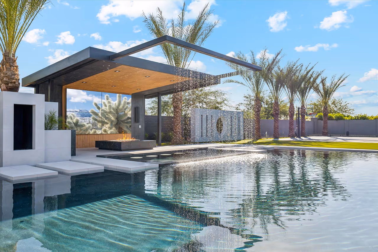 Modern backyard pool with a shaded lounge area featuring a fire pit and water jets creating an arc over the pool.