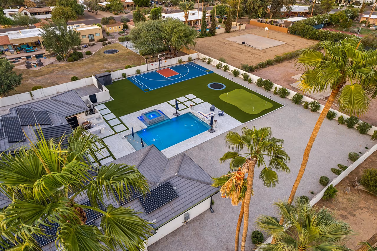 Aerial view of a modern backyard featuring a swimming pool with adjacent hot tub, a blue and red half basketball court, a putting green, trampoline, and palm trees.