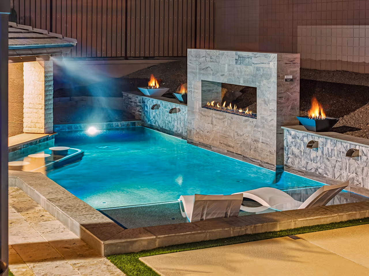 Modern backyard pool at night with built-in lounge chairs, three fire pits, and a stone fireplace feature.