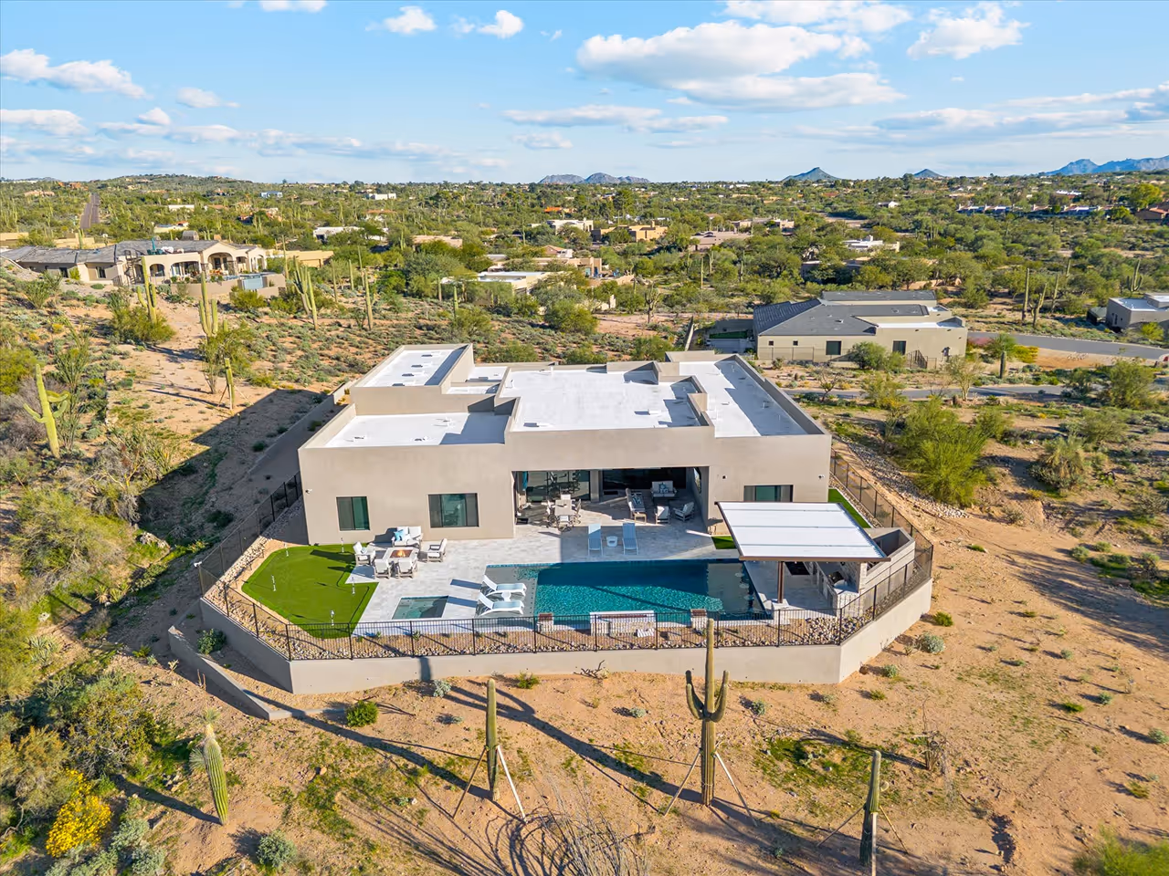 A modern single-story house with a flat roof, backyard pool, and patio, surrounded by desert landscape with cacti and scattered houses in the distance.