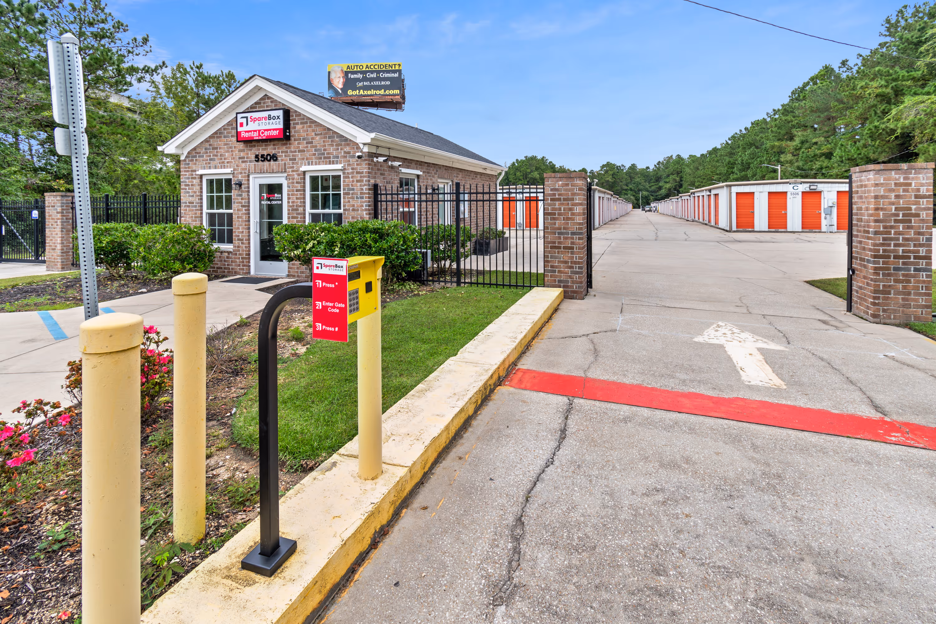 Entrance to SpareBox Storage with brick office, security gate, keypad, and rows of storage units with orange doors.
