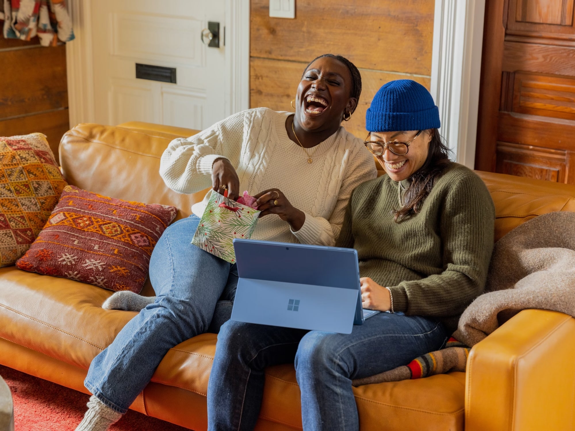 Two friends sitting on a tan leather couch, laughing together while looking at a laptop, one wearing a white sweater and the other a green sweater with a blue beanie.