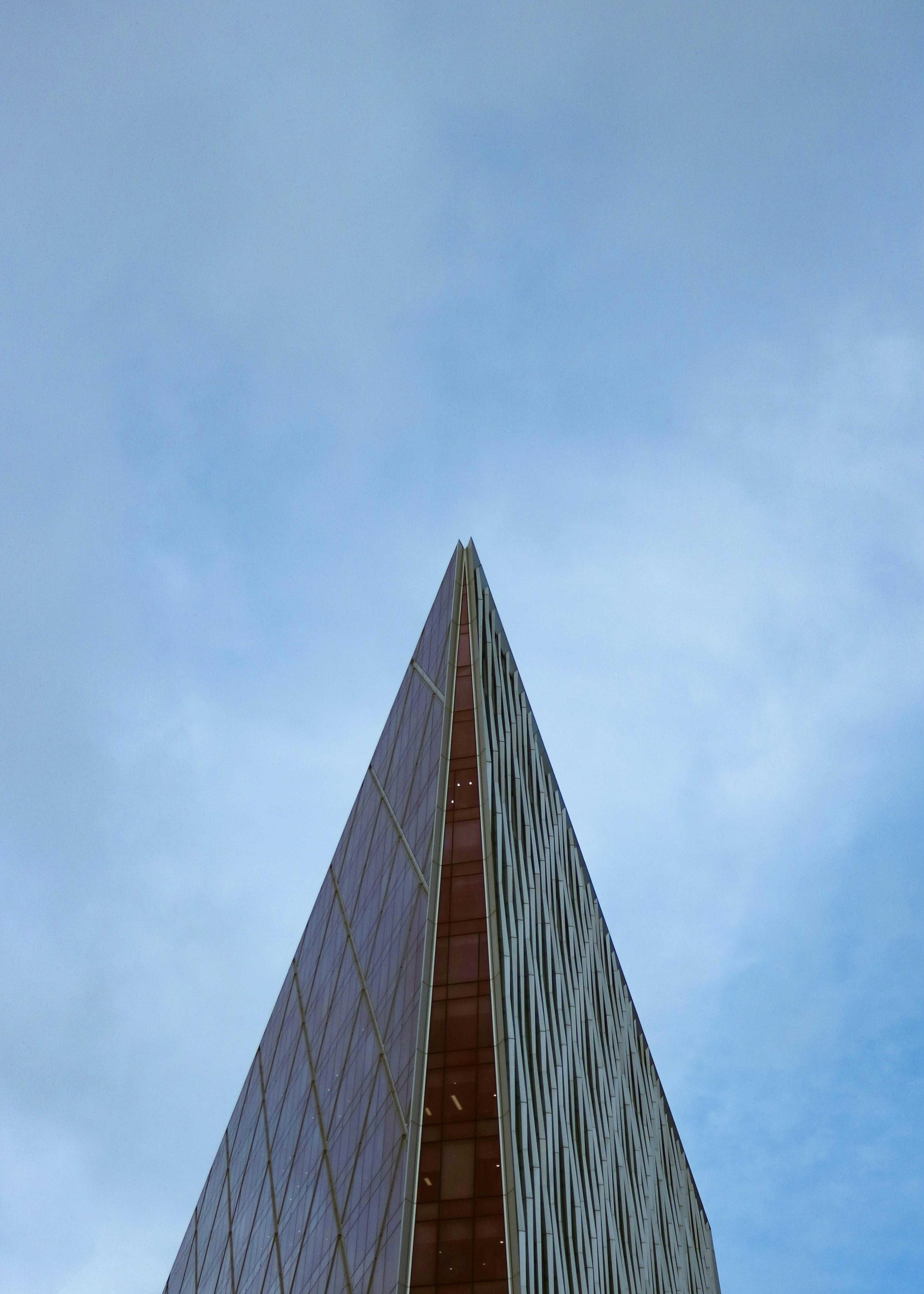 Sharp triangular peak of a modern glass and metal building against a cloudy blue sky.
