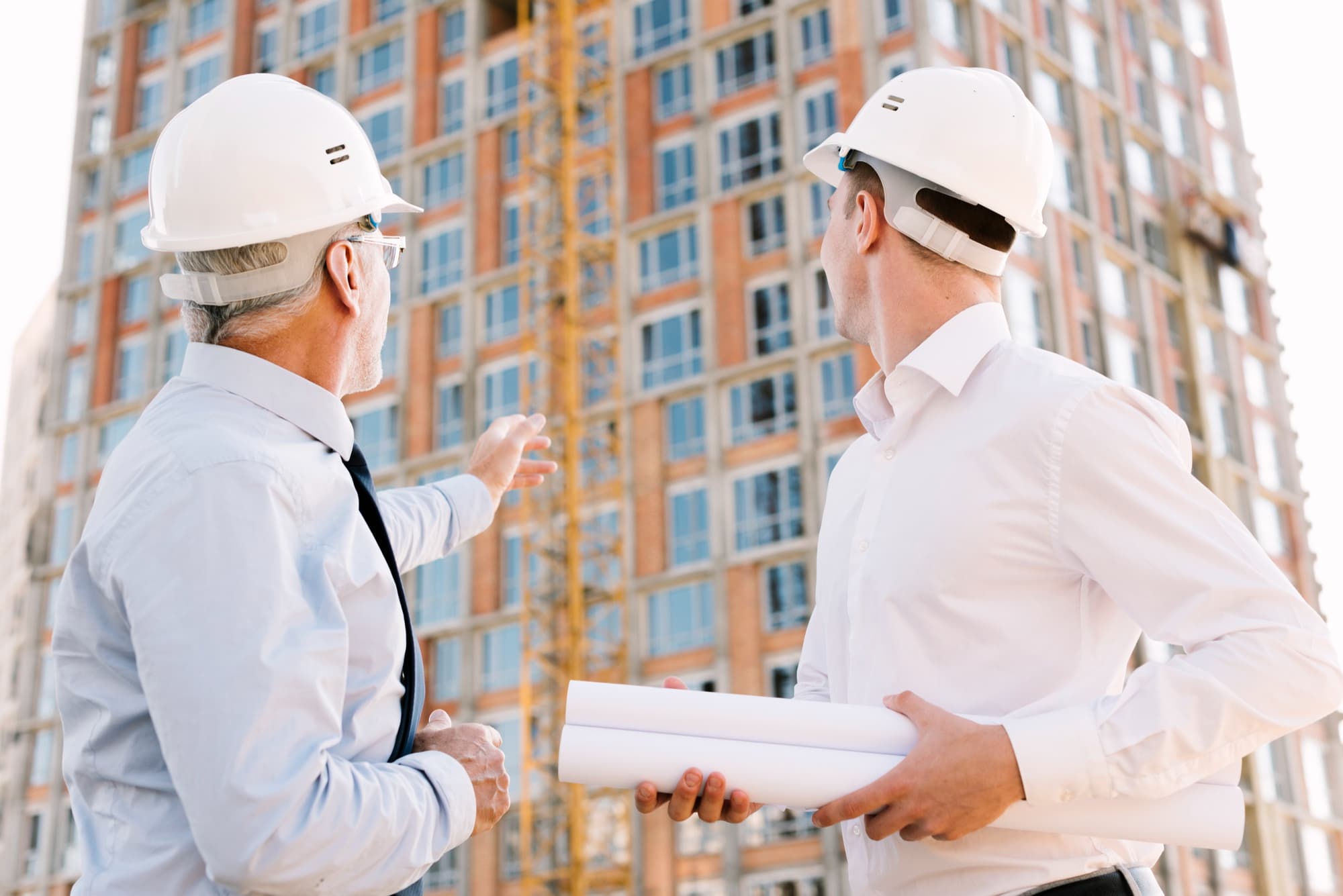 Two male construction engineers wearing white hard hats examining a building under construction, one holding blueprints.