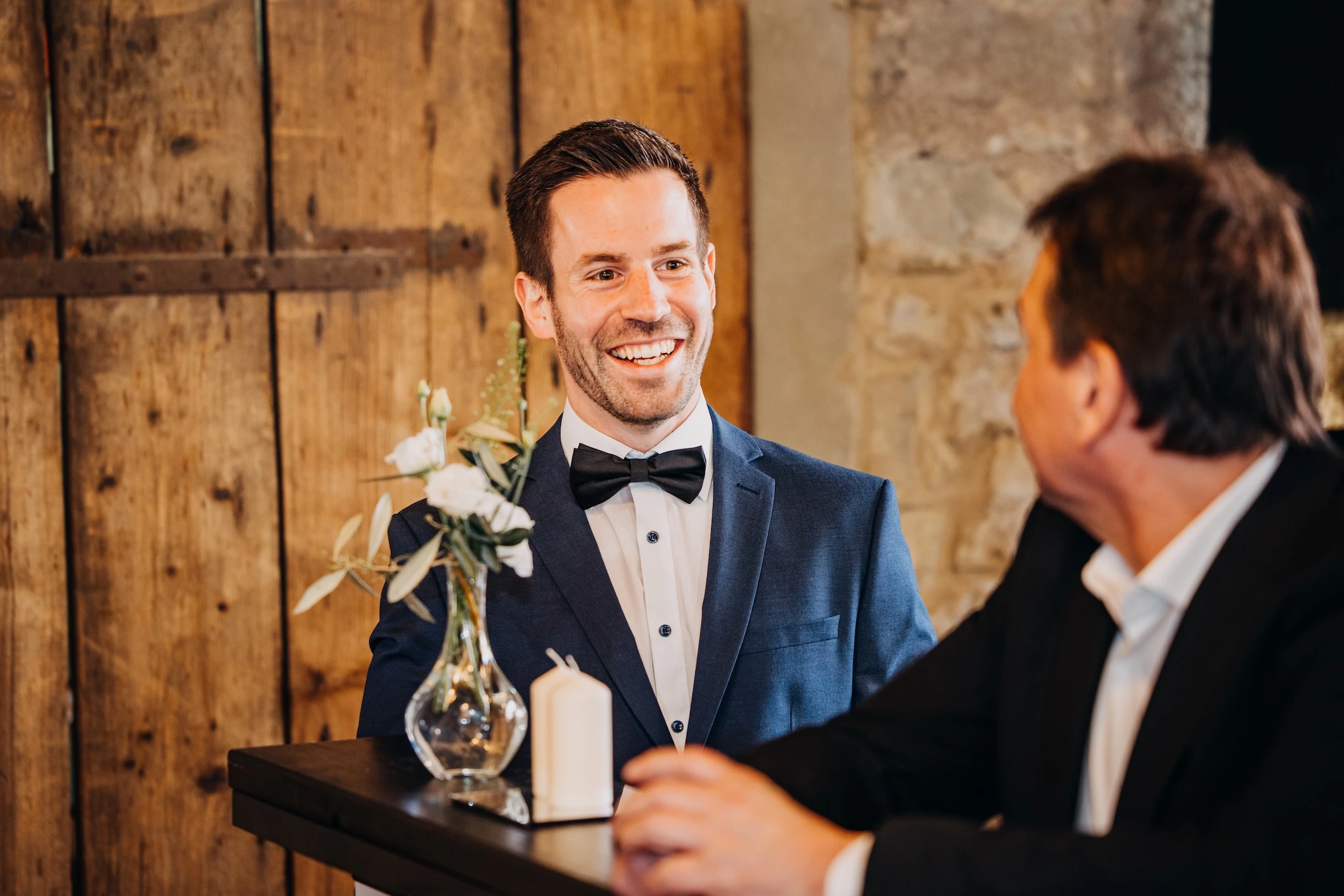 Smiling man in a dark blue suit and black bow tie sitting at a table with a small vase of white flowers and a candle, talking to another man in a black suit on a wedding ceremony