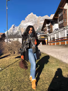 Woman in winter clothing standing on grass with snowy mountain and alpine buildings in the background under a clear blue sky.