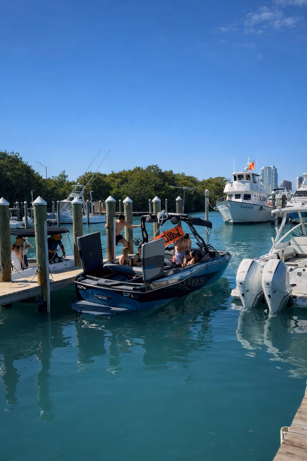 People on a sleek black wakeboard boat docked at a marina with clear blue water and other boats nearby under a clear sky.