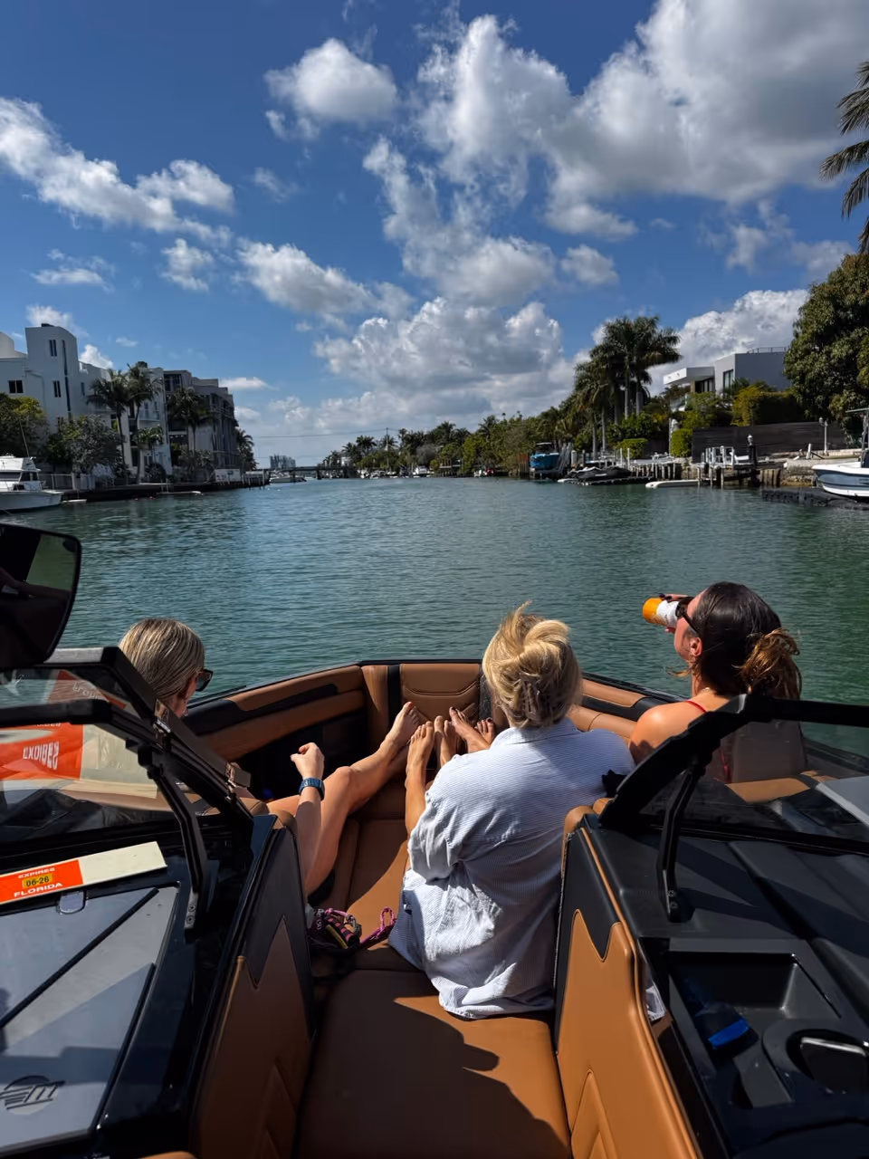 Three women relaxing on the bow of a boat cruising through a residential canal under a partly cloudy blue sky.