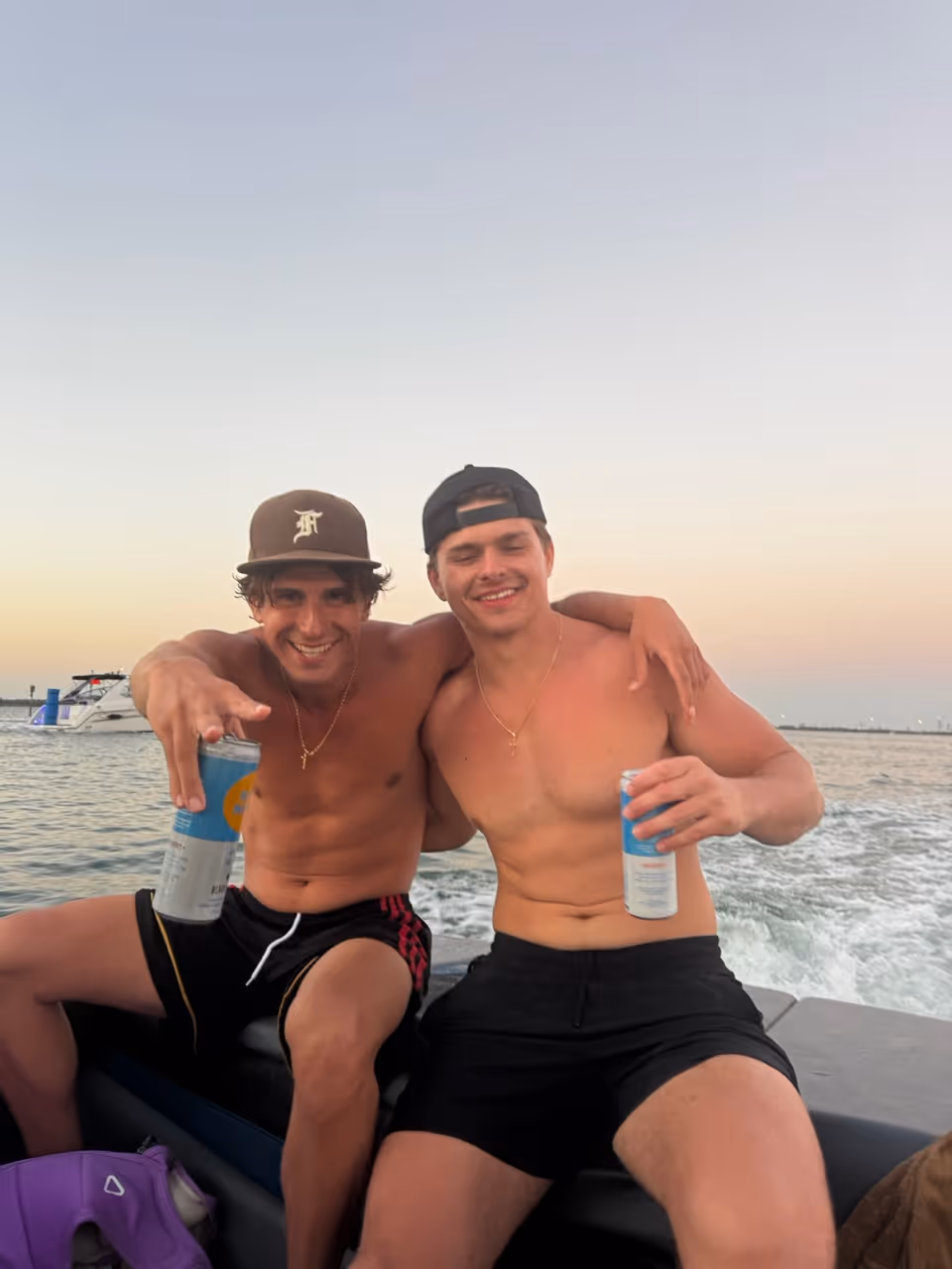 Two shirtless young men in shorts sitting on a boat, smiling, each holding a can, with water and another boat in the background at sunset.