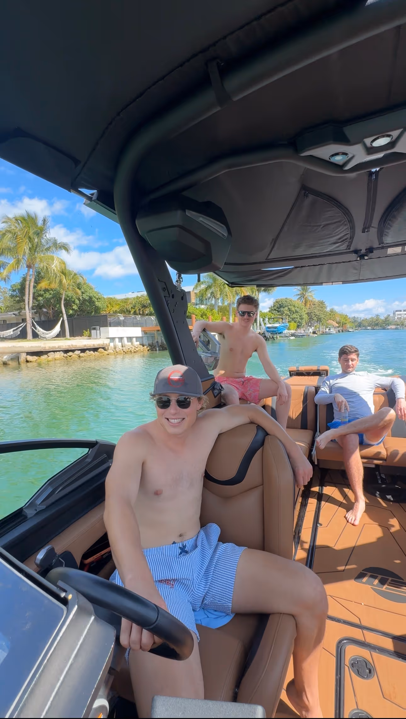 Three young men relaxing on a motorboat under a canopy on a sunny day with palm trees and calm water in the background.