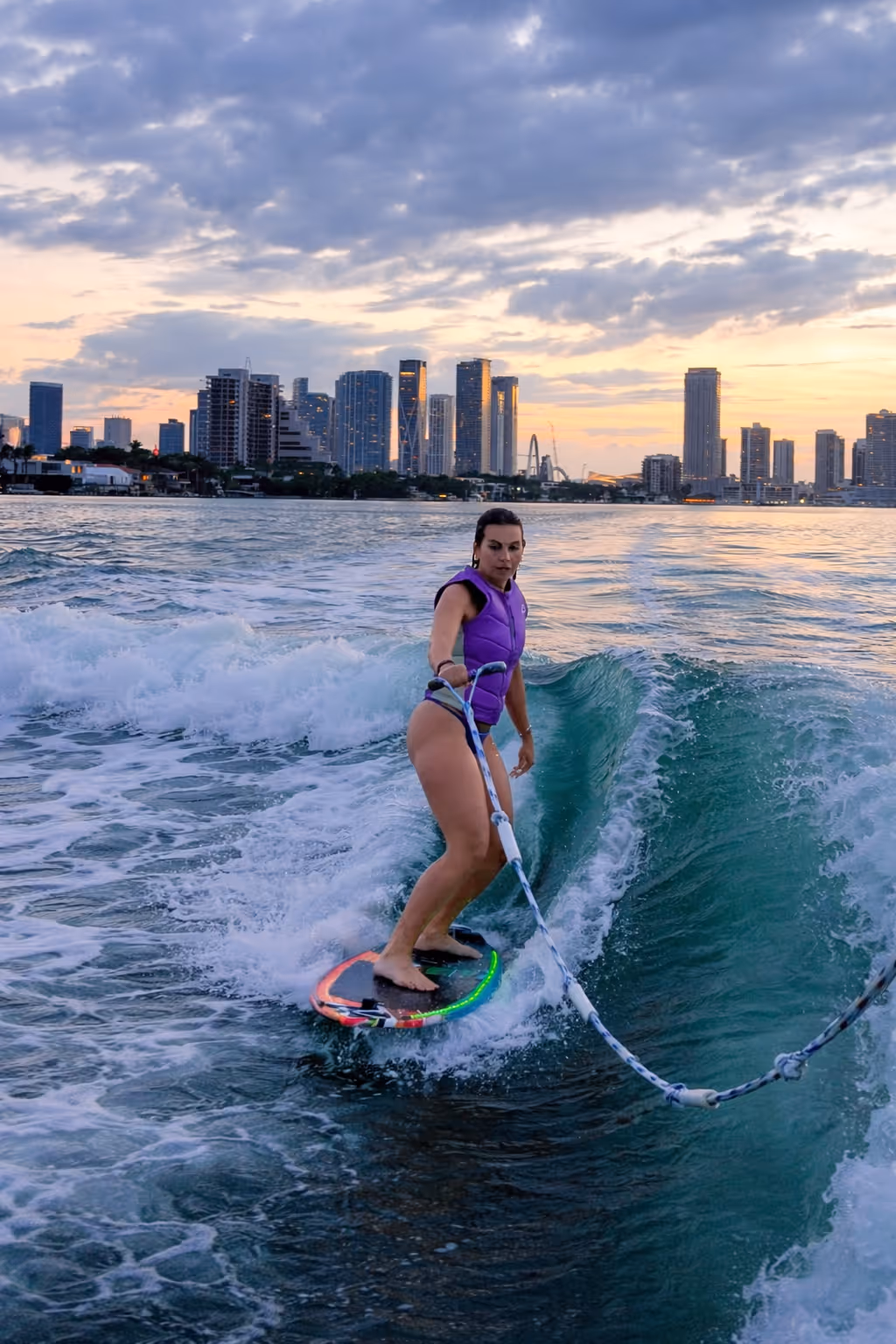 Mujer sentada descalza en un asiento de barco mirando el horizonte de Miami durante la puesta de sol con una tabla de wakeboard a su lado.