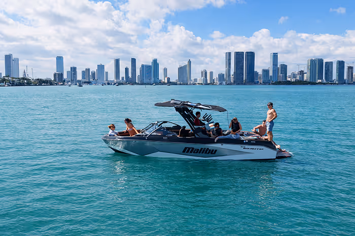 Group of people relaxing on a Malibu motorboat in calm blue water with Miami skyline under a partly cloudy sky.