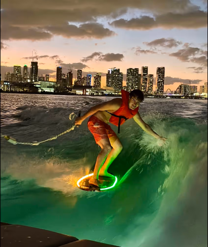 Man wearing a red life jacket wakesurfing at sunset with a city skyline in the background.