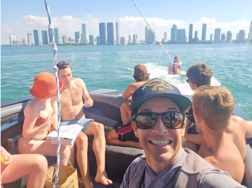 Group of six people on a boat with a city skyline in the background, one person taking a selfie while others look towards the water where someone is being towed on a wakeboard.