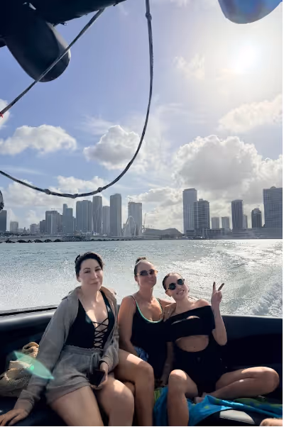 Three women sitting on a boat with a city skyline and water behind them under a bright sky with clouds.