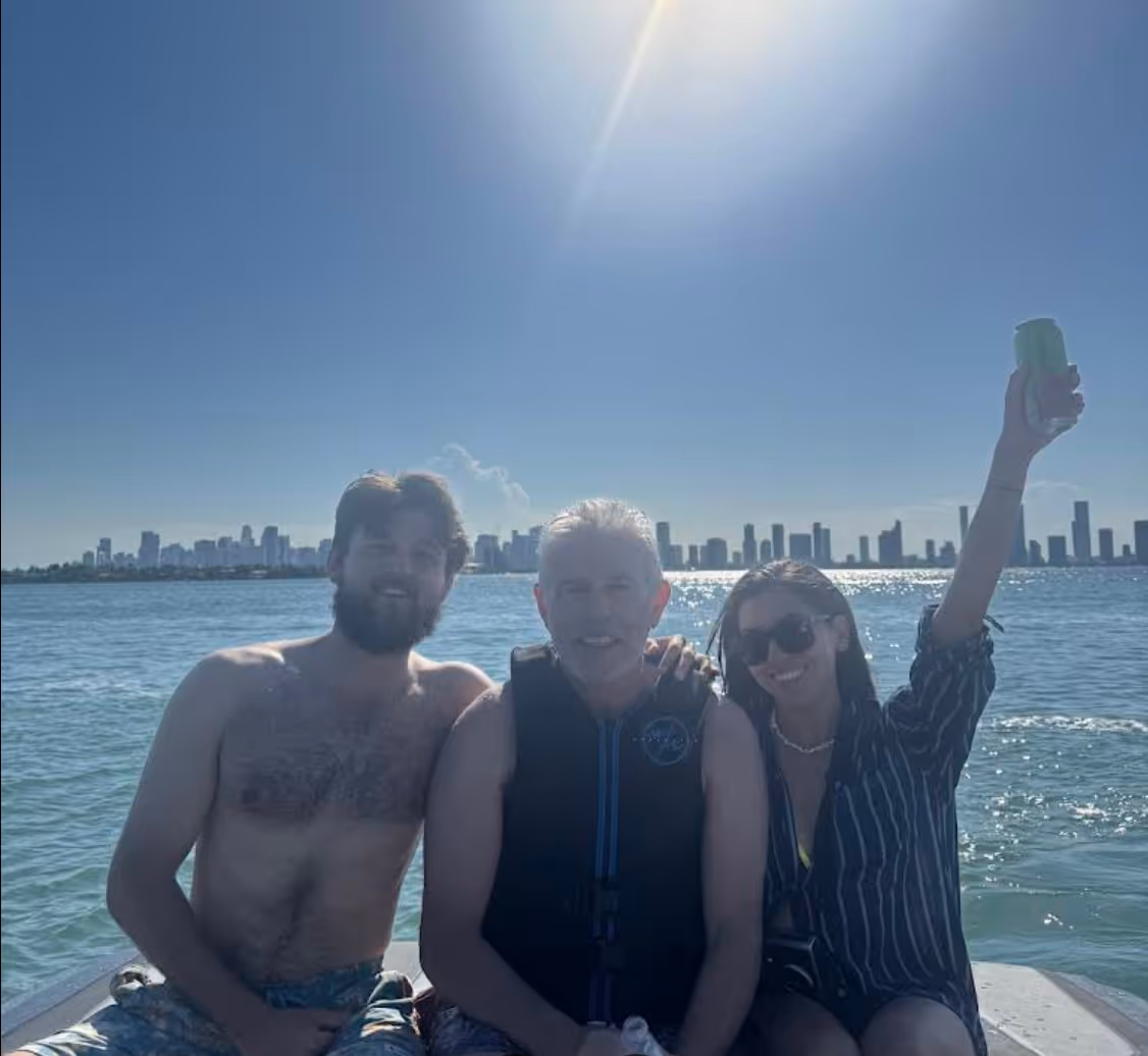 Three people on a boat with a city skyline and clear blue sky in the background, one person raising a drink.