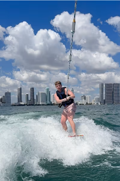 Person wakeboarding on water with city skyline and cloudy blue sky in the background.