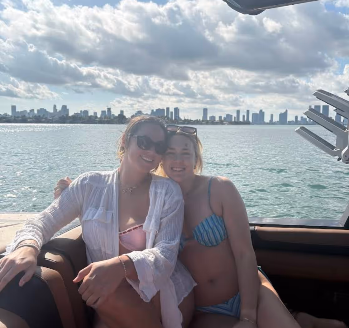 Two smiling women in swimsuits sitting together on a boat with a city skyline and partly cloudy sky in the background.