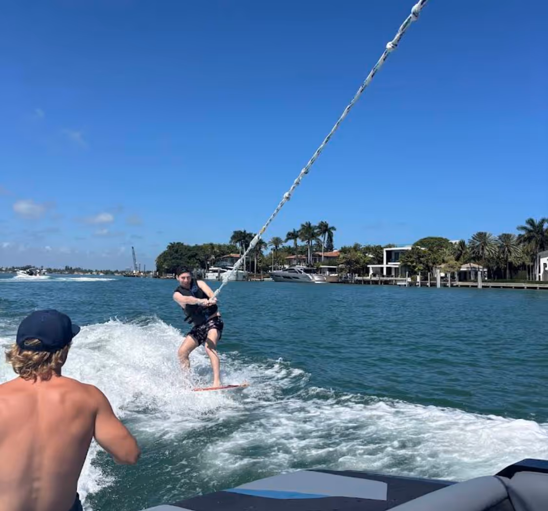 Person wakeboarding behind a boat on a sunny day with waterfront houses and palm trees in the background.