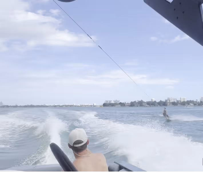 Vista desde un barco en movimiento que muestra a una persona haciendo wakeboard en un cuerpo de agua tranquilo bajo un cielo parcialmente nublado.