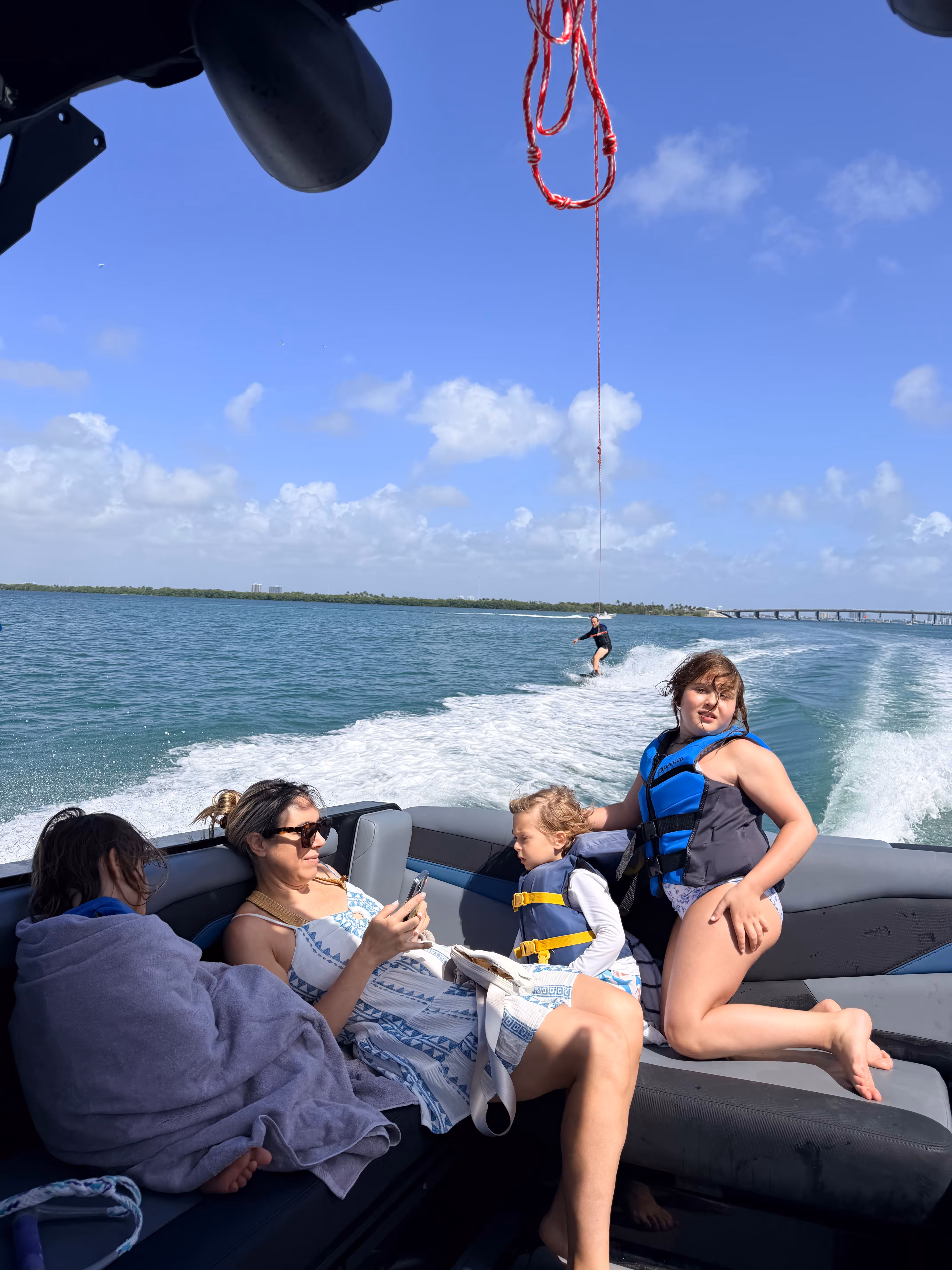 Mujer y tres niños en un barco, con un niño haciendo wakeboard detrás en aguas azules bajo un cielo despejado.