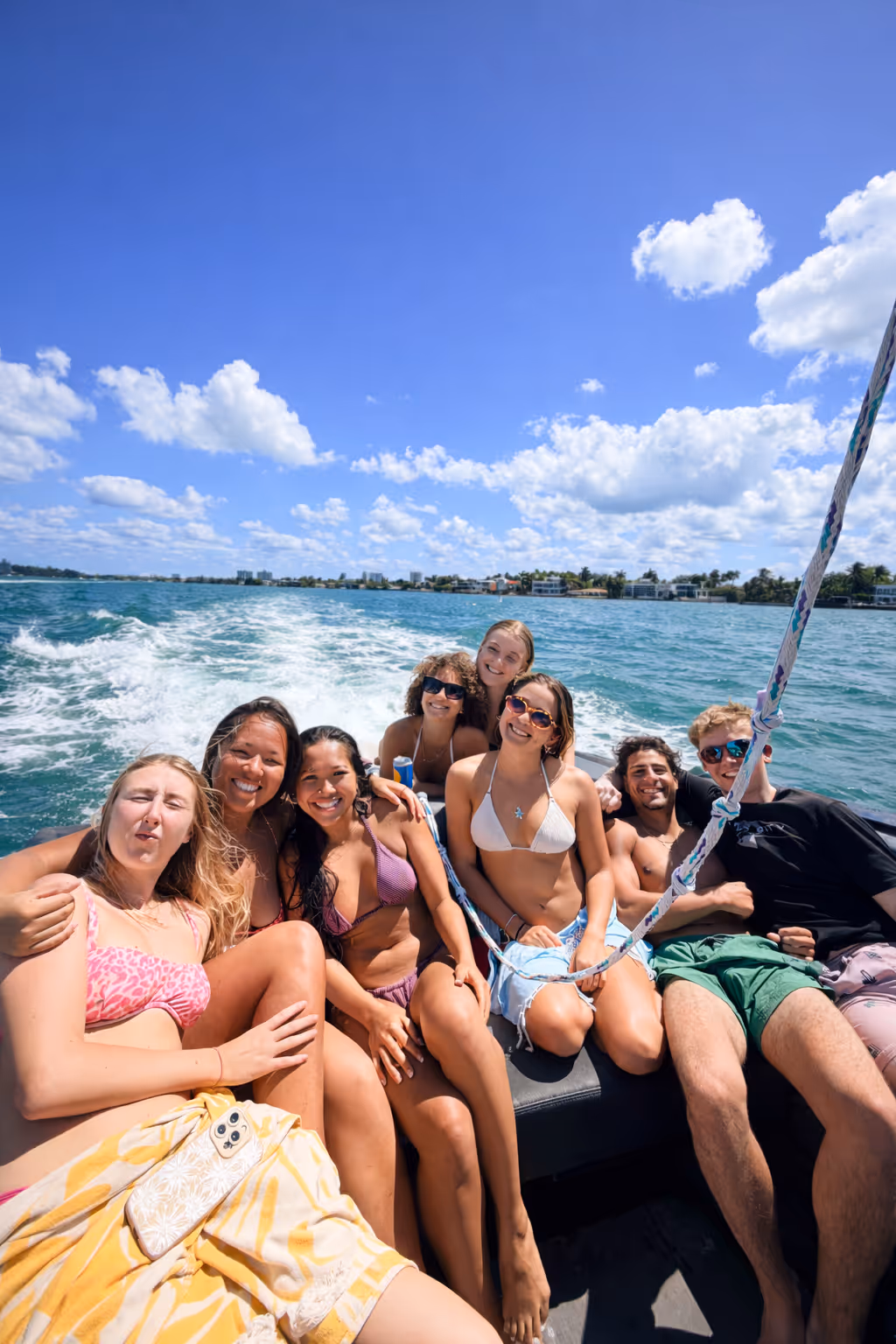 Group of seven friends smiling and relaxing in swimsuits on a boat cruising on clear blue ocean water under a sunny sky with scattered clouds.