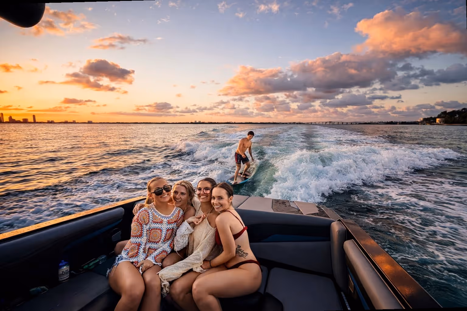 Four women smiling and sitting closely on a boat while a man surfboards on the water behind them during a colorful sunset.