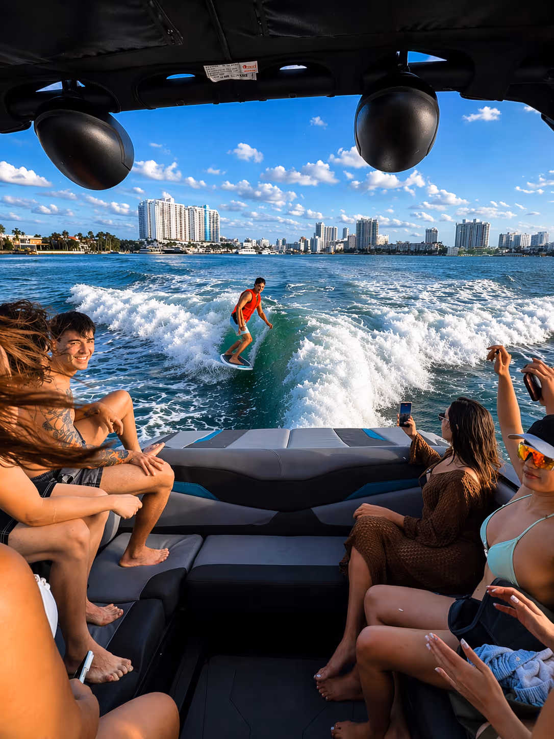 A group of people on a boat watching a wakesurfer riding the wave behind the boat in a city waterfront setting under blue sky.