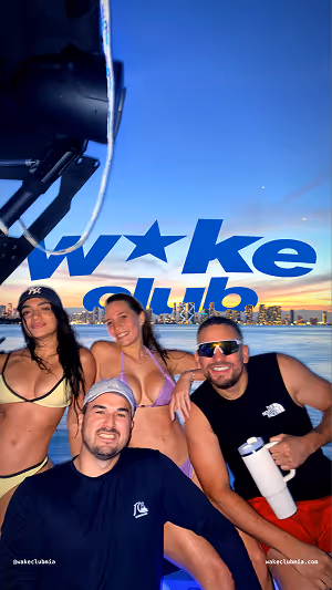 Two women smiling and sitting closely together on a boat with a city skyline and water in the background under a partly cloudy sky.