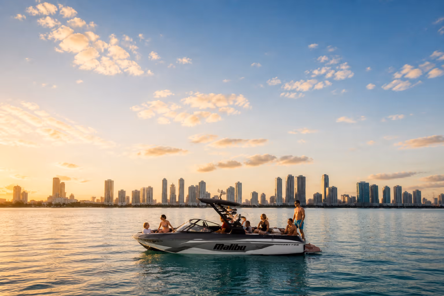 Group of people relaxing on a Malibu boat in calm water with city skyline at sunset.