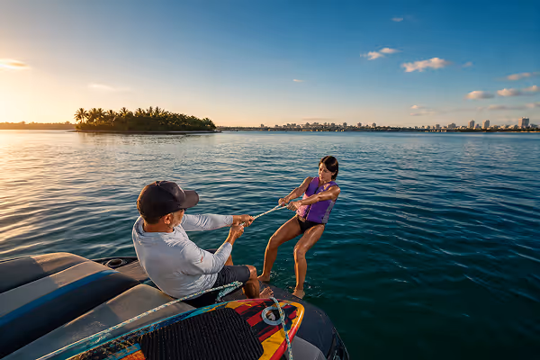Man in a cap holding a rope and helping a woman in a purple life jacket emerge from water near a boat during a sunset with an island and city skyline in the background.
